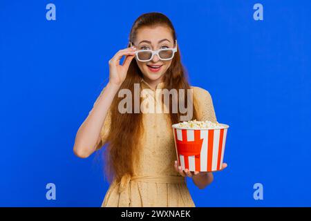 Begeisterte junge Erwachsene Frau in 3D-Brille isst Popcorn und sieht interessante fernsehserien, Sportspiele, Filme und Online-Inhalte von sozialen Medien an. Rothaariges Mädchen isoliert auf blauem Atelierhintergrund drinnen Stockfoto