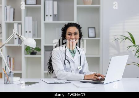 Eine fröhliche Latino-Ärztin in einem weißen Mantel lächelt, während sie in einem modernen Büro an ihrem Laptop arbeitet. Stockfoto