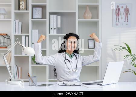 Eine fröhliche latino-Ärztin mit lockigem Haar hebt ihre Arme in triumphaler Pose und feiert einen professionellen Erfolg in ihrer Arztpraxis. Stockfoto