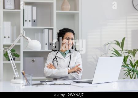 Porträt einer fröhlichen hispanischen Ärztin mit Brille, die mit einem Laptop in ihrem Klinikbüro sitzt und Professionalität und Pflege ausstrahlt. Stockfoto