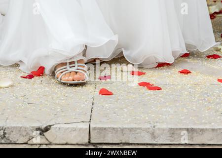 Der Fuß einer Braut, der mit einer schicken Sandale verziert ist, tritt zart auf eine romantische Spur aus Rosenblättern und Konfetti, die sie den Gang entlang markiert Stockfoto