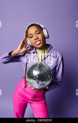 Glückliche junge afroamerikanische Frau in stilvoller Kleidung und kabellosen Kopfhörern, die Discokugel halten Stockfoto