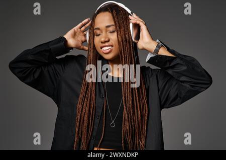 Fröhliche dunkelhäutige Frau mit Dreadlocks, die Musik in kabellosen Kopfhörern auf Grau hört Stockfoto