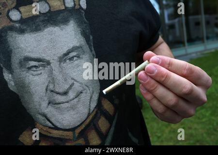 München, Deutschland. April 2024. Ein Teilnehmer einer unter dem Motto „Liberalitas Bavariae statt CSU-Verbotsirtum“ registrierten Kundgebung hält vor einem T-Shirt mit einem Porträt des bayerischen Ministerpräsidenten Markus Söder (CSU) im CSU-Staatsleitungshaus einen Joint. Quelle: Karl-Josef Hildenbrand/dpa/Alamy Live News Stockfoto