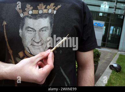 München, Deutschland. April 2024. Ein Teilnehmer einer unter dem Motto „Liberalitas Bavariae statt CSU-Verbotsirtum“ registrierten Kundgebung hält vor einem T-Shirt mit einem Porträt des bayerischen Ministerpräsidenten Markus Söder (CSU) im CSU-Staatsleitungshaus einen Joint. Quelle: Karl-Josef Hildenbrand/dpa/Alamy Live News Stockfoto