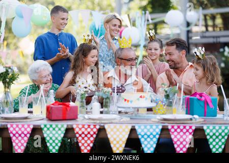 Gartengeburtstag für ältere Männer. Hübscher älterer Geburtstagsmann, der Kuchen von der Familie bekommt. Stockfoto
