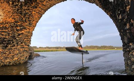 Tragflächenreiter gleitet mit seinem Brett in einem der Kanäle der Ria de Aveiro in Portugal während des Sonnenuntergangs über dem Wasser. Stockfoto