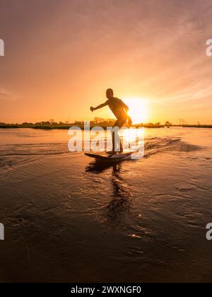 Tragflächenreiter gleitet mit seinem Brett in einem der Kanäle der Ria de Aveiro in Portugal während des Sonnenuntergangs über dem Wasser. Stockfoto