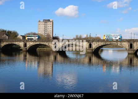 Doppeldeckerbusse mit Postkutsche überqueren die historische Skerton Bridge über den Fluss Lune in Lancaster, Lancashire, England bei Flut am 30. März 2024. Stockfoto