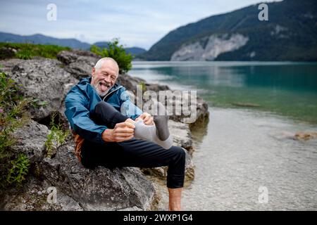 Aktiver älterer Mann, der sich Socken anzieht nach Barfußspaziergang im kalten Wasser im Bergsee. Senioren genießen die Natur im Frühjahr. Stockfoto