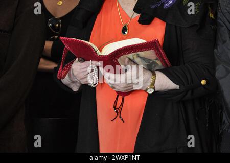 Eine ältere Frau, in einer orangen Bluse, schwarzem Mantel, goldenen Uhr am Handgelenk, hält eine Bibel und einen weißen Rosenkranz in den Händen. Stockfoto