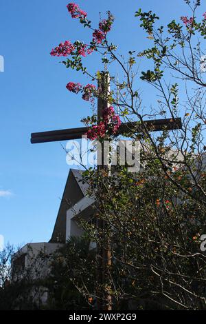 Ein Holzkreuz zwischen den grünen Blättern eines Baumes mit roten, rosa und gelben Blüten. Im Hintergrund, Teil einer Kirche unter einem wunderschönen blauen Himmel. Stockfoto