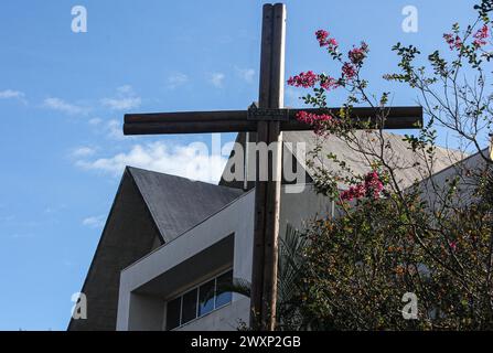 Ein Holzkreuz zwischen den grünen Blättern eines Baumes mit roten, rosa und gelben Blüten. Im Hintergrund, Teil einer Kirche unter einem wunderschönen blauen Himmel. Stockfoto