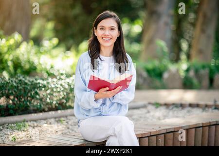 Ein enthusiastischer junger Leser sitzt mit einem strahlenden Lächeln auf einer Parkbank, vertieft in ein fesselndes rotes Buch Stockfoto