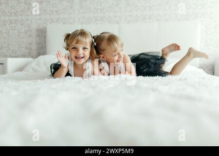 Zwei junge Mädchen, die elegante schwarze Kleider tragen, liegen auf dem Bauch, stehen einander mit einem Lächeln auf einer flauschigen weißen Bettdecke gegenüber und spielen mit Stockfoto