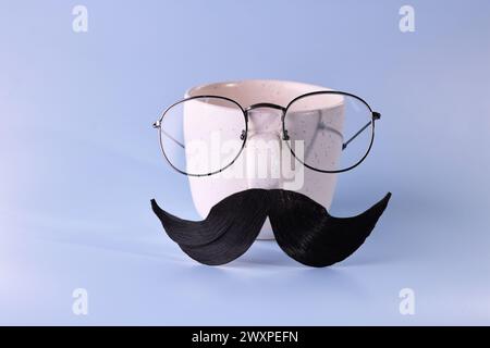Männergesicht aus künstlichem Schnurrbart, Brille und Tasse auf hellblauem Hintergrund Stockfoto