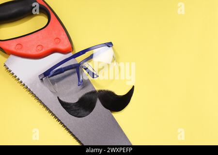 Männergesicht aus künstlichem Schnurrbart, Schutzbrille und Handsäge auf gelbem Hintergrund, Draufsicht. Leerzeichen für Text Stockfoto