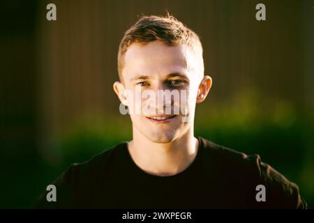 Männliches Außenporträt in Abendbeleuchtung, Nahaufnahme Gesicht eines rauen weißen Mannes in seinen 20ern, der in die Kamera vor verschwommener ländlicher Landschaft blickt. Stockfoto