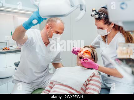 Zahnärztlicher Klinikpatiententermin. Zahnarztfrau in Lupenbrille, die Zahnprophylaxe mit medizinischen Werkzeugen macht. Junger Mann Assistent, der Licht zeigt. Stockfoto
