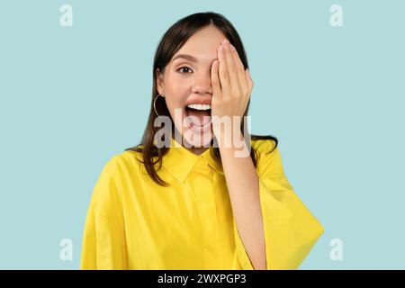 Glückliche überraschte Frau, die ein Auge mit einer Hand bedeckt, auf blauem Studiohintergrund Stockfoto