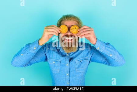 Ein lächelnder bärtiger Mann hat die Augen mit Haferflockenkeksen bedeckt. Leckerer süßer Snack. Gutaussehender Mann mit Schokoladenkeksen Kekse. Frühstück oder Mittagessen. Attraktiv Stockfoto