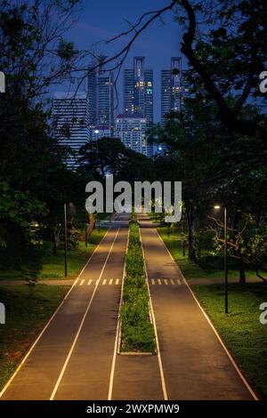 Beleuchtete Wolkenkratzer hinter einem Pfad oder Laufbahn und üppige Bäume im Benjakitti Forest Park in Bangkok, Thailand bei Dämmerung. Stockfoto