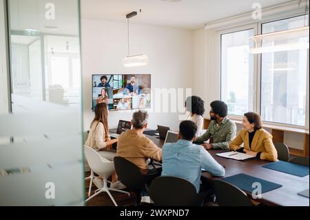 Ein Team arbeitet aktiv mit Kollegen aus der Ferne über einen großen Bildschirm zusammen und veranschaulicht das moderne hybride Arbeitsmodell, bei dem Technologie die Lücke zwischen Teammitgliedern im Büro und Remote-Mitarbeitern überbrückt Stockfoto