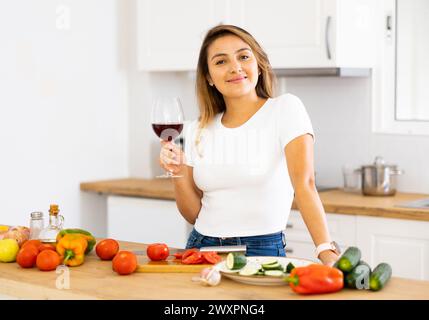 Fröhliche junge hispanische Frau, die während der Zubereitung des Abendessens Wein genießt Stockfoto
