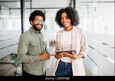 Zwei Kollegen stehen in einem modernen Büro nebeneinander, zeigen ein Gefühl der Partnerschaft, die Frau hält ein Tablet, teilt eine neue Idee oder ein Projekt, beide lächeln, was auf eine positive Zusammenarbeit hinweist Stockfoto