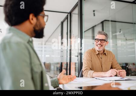 Zwei männliche Berufskollegen führen eine freundliche Diskussion im Büro, der ältere Mann mit Brille zeigt ein warmes Lächeln, was auf ein positives Arbeitsumfeld hinweist Stockfoto