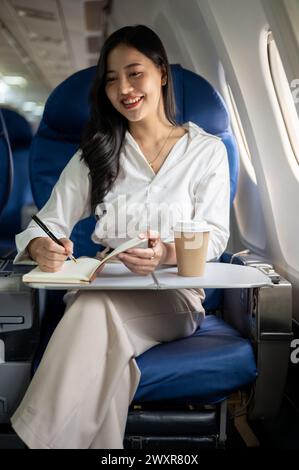 Eine attraktive asiatische Geschäftsfrau macht Notizen in ihrem Notizbuch, während sie zu einem Geschäftstreffen in eine andere Stadt fliegt. Eine Geschäftsfrau ist im Flugzeug Stockfoto