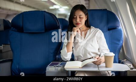 Eine elegante asiatische Geschäftsfrau konzentriert sich auf ihre Arbeit und überprüft ihren Terminplan in ihrem Notizbuch, während sie zu einem Geschäftstreffen in eine andere Stadt fliegt Stockfoto