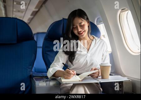 Eine attraktive asiatische Geschäftsfrau macht Notizen in ihrem Notizbuch, während sie zu einem Geschäftstreffen in eine andere Stadt fliegt. Eine Geschäftsfrau ist im Flugzeug Stockfoto