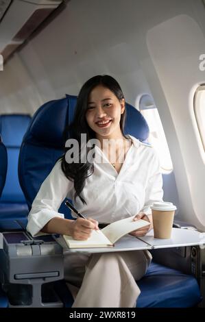Eine attraktive asiatische Geschäftsfrau macht Notizen in ihrem Notizbuch, während sie zu einem Geschäftstreffen in eine andere Stadt fliegt. Eine Geschäftsfrau ist im Flugzeug Stockfoto