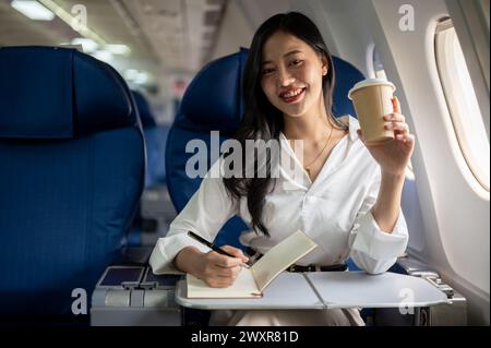 Eine schöne asiatische Geschäftsfrau lächelt in die Kamera, während sie im Flugzeug an einem Fenstersitz sitzt und geschäftlich unterwegs ist. Geschäftsleute und t Stockfoto