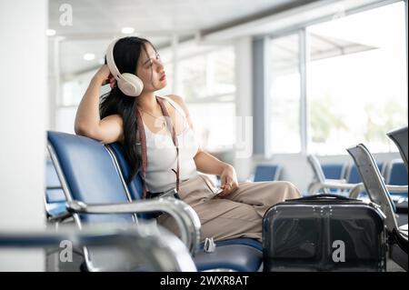 Eine asiatische Passagierin fühlt sich müde und gelangweilt, während sie im Flughafenterminal auf ihren Anruf wartet und Musik über ihre Kopfhörer hört Stockfoto