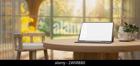 Ein digitales Tablet mit weißem Bildschirm auf einem Holztisch in einem komfortablen, neutralen Wohnzimmer mit einem großen Fenster mit Blick auf einen wunderschönen Garten auf einem Su Stockfoto