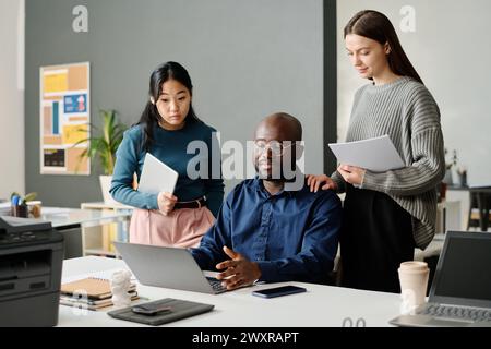 Junger afroamerikanischer Manager, der am Tisch vor dem Laptop sitzt und weibliche Kollegen um Rat zu seinem Präsentationsdesign bittet Stockfoto