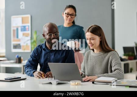 Gruppe von drei jungen ethnisch unterschiedlichen Menschen, die im Büro arbeiten, sich den Laptop-Bildschirm ansehen und die Geschäftsstrategie diskutieren Stockfoto