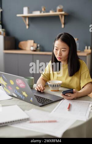 Vertikale Aufnahme einer jungen asiatischen Universitätsstudentin, die morgens beim Frühstück am Tisch sitzt und einen Laptop benutzt Stockfoto