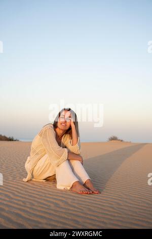 Lächelnde junge barfuß-Frau in legeren Kleidern, die in die Kamera blickt, während sie am Abend allein mit Schatten auf den Dünen von Corralejo gegen blaue s sitzt Stockfoto