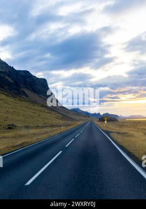 Eine lange Straße ohne Autos und einen Berg im Hintergrund. Der Himmel ist bewölkt und die Sonne untergeht Stockfoto