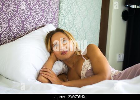 Friedliche junge Frau liegt im Bett, legt ihren Kopf auf ein Kissen und blickt nachdenklich Stockfoto