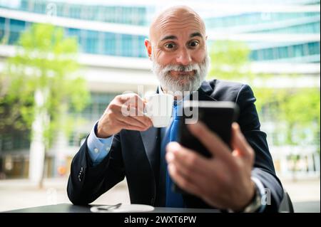 Senior stylischer Manager mit weißem Bart, der draußen einen Kaffee trinkt, während er sein Smartphone mit einem schockierten Ausdruck benutzt Stockfoto
