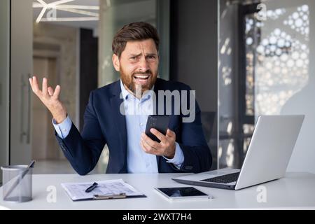Porträt eines wütenden jungen Geschäftsmannes, der an einem Schreibtisch im Büro sitzt, ein Telefon in der Hand hält und enttäuscht von der Kamera aussieht. Stockfoto