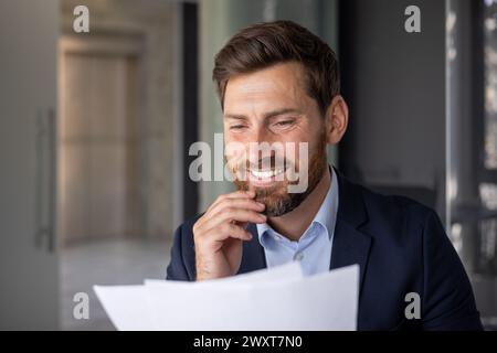 Ein lächelnder Geschäftsmann im Anzug untersucht Papiere und zeigt Selbstvertrauen und Professionalität in einem gut beleuchteten Büro. Stockfoto