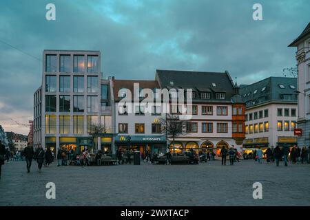 Eine Gruppe von Menschen, die auf einem Stadtplatz spazieren, der von Gebäuden gesäumt ist, unter einem bewölkten Himmel. Der Asphaltpflaster steht im Kontrast zu den grünen Bäumen und Wasserspielen in der Nähe Stockfoto