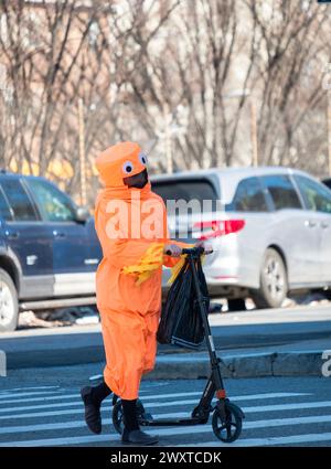 Auf purim 2024 geht ein orthodoxer jüdischer Junge mit seinem Roller in einem orangen Ganzkörperkostüm über die Straße. Auf der Lee Avenue in Williamsburg, Brooklyn, NYC. Stockfoto