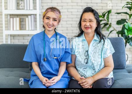 Eine Krankenschwester und eine asiatische Dame sitzen auf dem Sofa und schauen in die Kamera Stockfoto