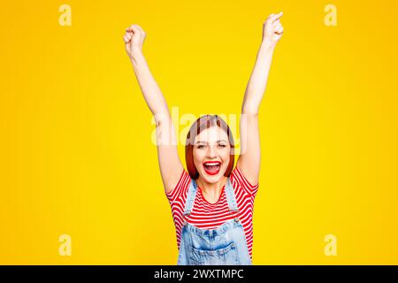 Porträt eines netten, lebendigen, rothaarigen, fröhlichen, lächelnden, jungen Mädchens mit offenem Mund, erhobenen Händen, isoliert auf blauem Hintergrund Stockfoto